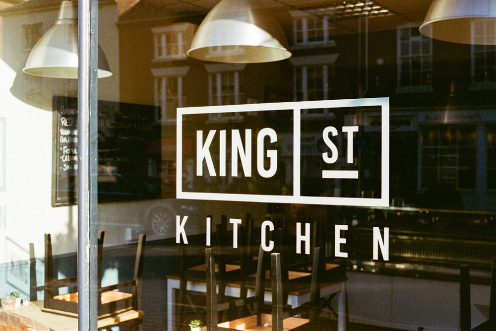 The window of a closed cafe with the words ‘King St Kitchen’ with chairs stacked on tables in the evening sunlight inside.