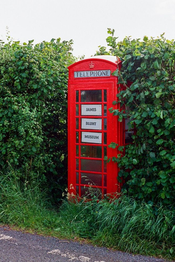 A bright red telephone box surrounded by a tall green hedge with a sign saying ‘James Blunt Musuem’.