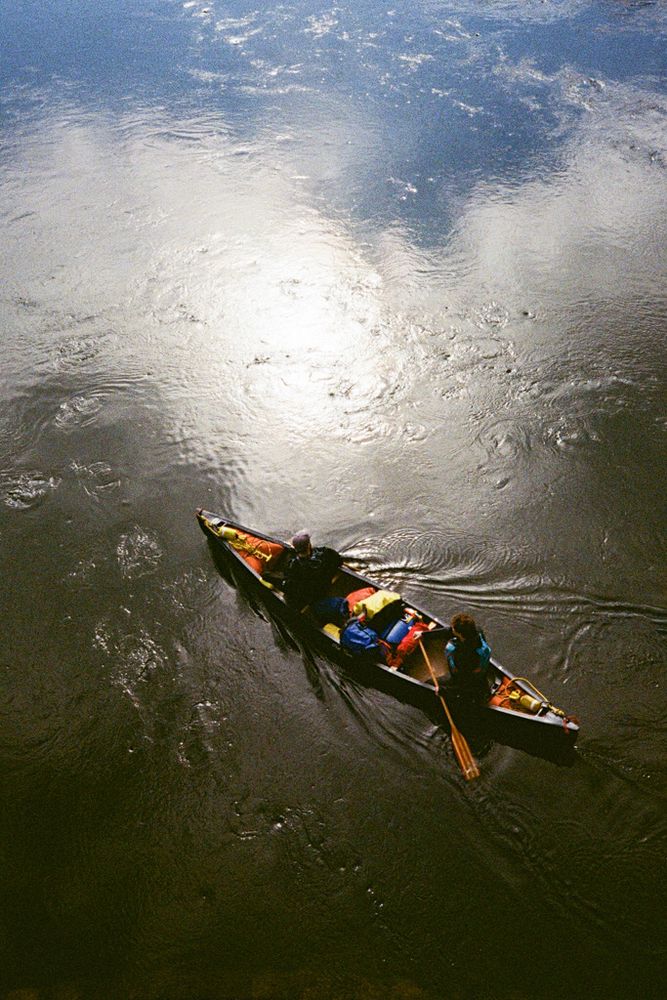 Looking down from a bridge as a canoe with two people paddling drifts through the darkly-coloured water of a river, with the bright reflection of the sun in the water’s surface.