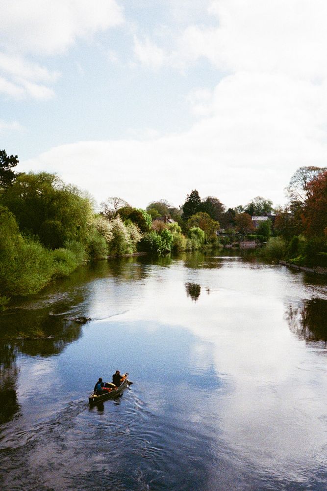 A view from a bridge looking down on a river as it flows into the distance. A canoe with two people in it drift downstream with green trees on either bank.