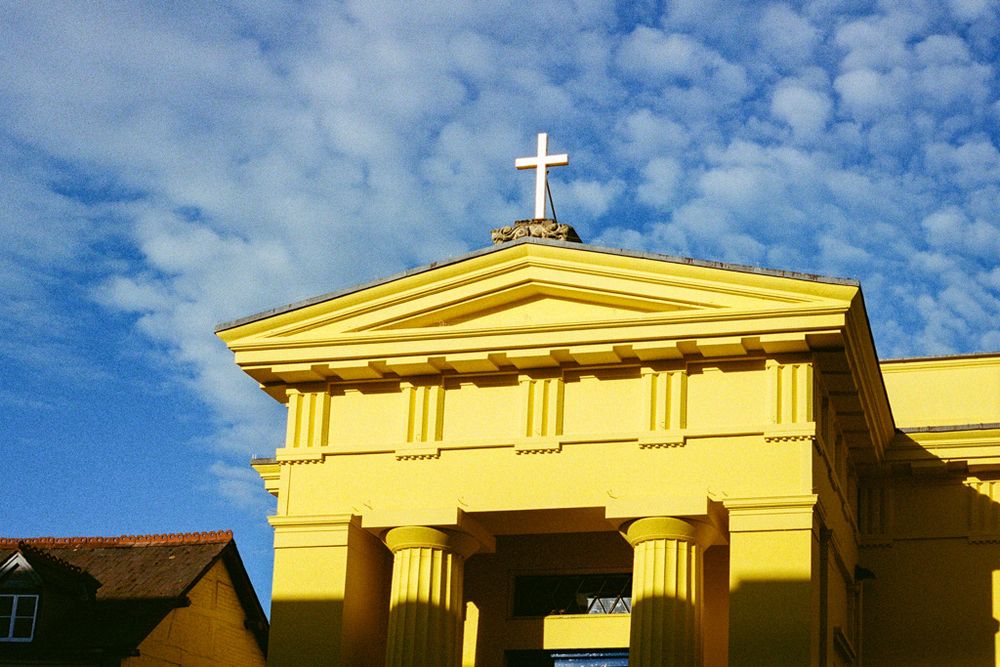 Looking up at a brightly painted yellow church building in evening sunlight, with a golden cross on the top & blue sky with light clouds behind.