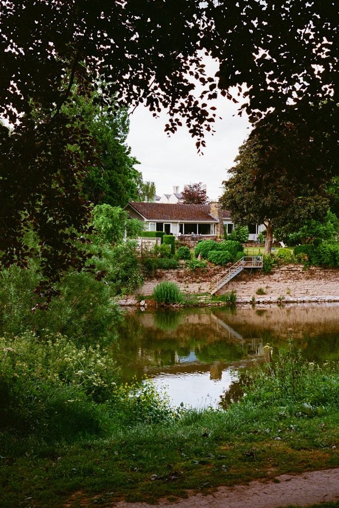A view looking across a river framed by trees above & green grass in the foreground. On the other side of the river there is a building with white windows that is reflected in the water.