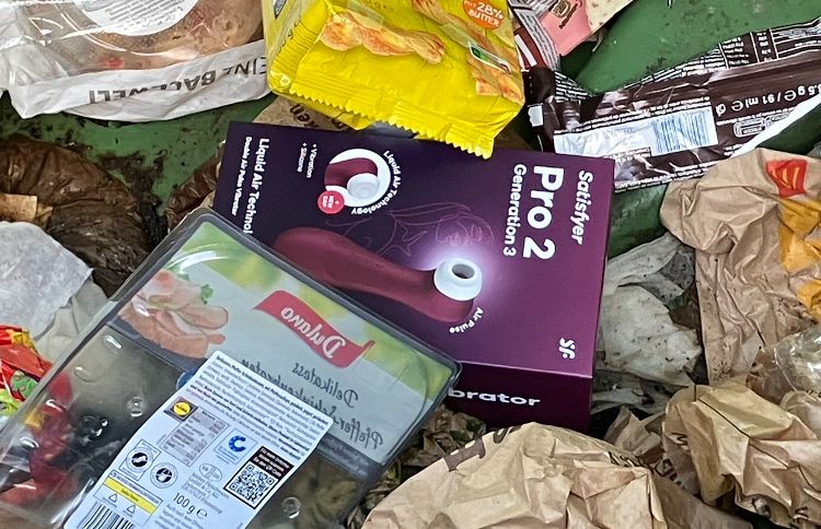 Aufnahme des Mülls in einer großen Mülltonne. Mittig liegt die Verpackung eines Satisfyers Pro2, zwischen anderem Müll, u.a. braune Papiertüten, eine Schnittwurstpackung. 