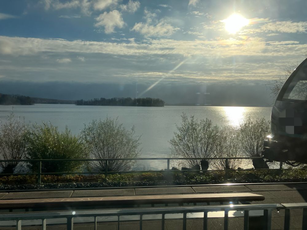 Blick aus einem Zugfenster. Im Vordergrund des nebenliegende Bahnhofsgleis, rechts ragt die Spitze eines Zuges ins Bild. Hinter der Bahnstrecke ein paar Büsche und Bäume, dann ein See, der glatt und grau, durchbrochen von Lichtreflexen, daliegt. Mittig im See eine kleine, baumbestandene Insel, am hinteren Ufer ebenfalls Begrünung. Der Himmel ist blau mit Wolken, aber die Sonne blendet so hell, dass die Farben ganz blass sind. 