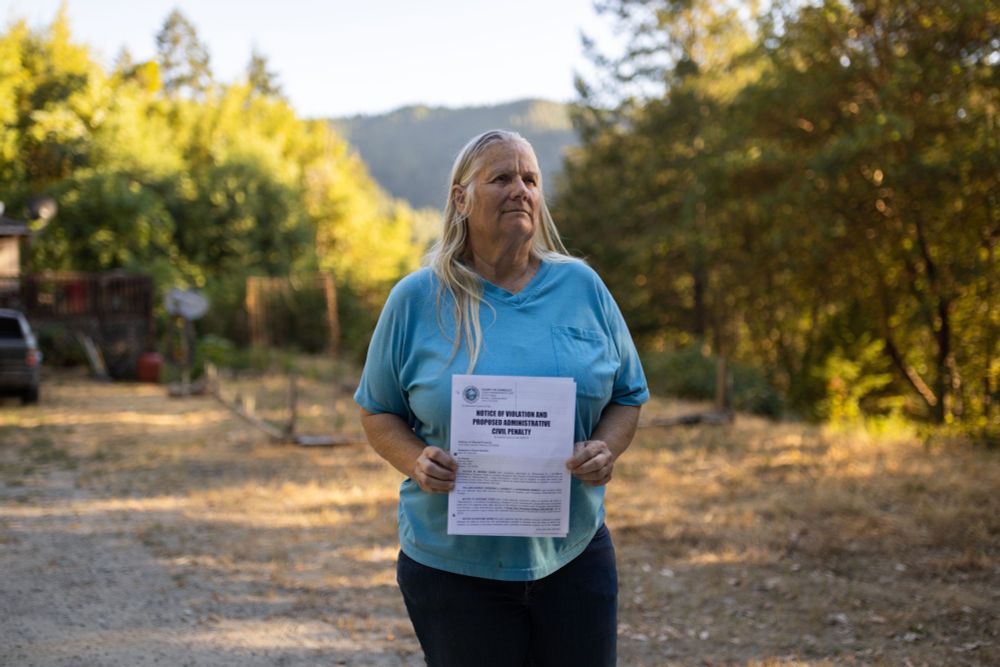 Rhonda Olson standing on her mountainous property in the northwest corner of Humboldt County, CA.  She's holding a Notice of Violation that charged her with millions in fines for someone else's conduct.
