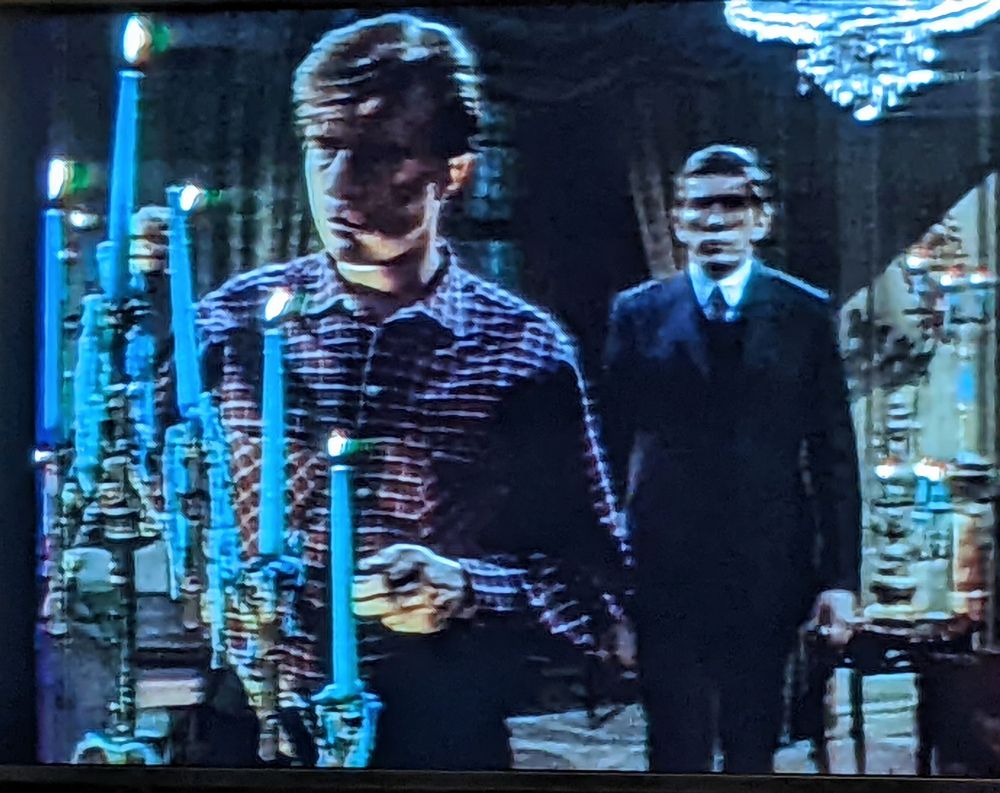 Man in red checked shirt lighting fresh blue candles in a candelabra while a stern faced man in black suit stands behind him.
