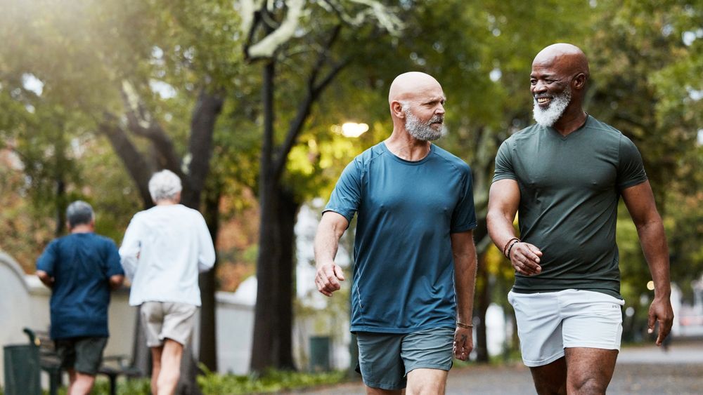 Two men walking in a park. They are wearing athletic clothing and appear to be engaged in conversation. In the background, two other people are also walking or jogging on the path. The park is filled with trees and greenery.