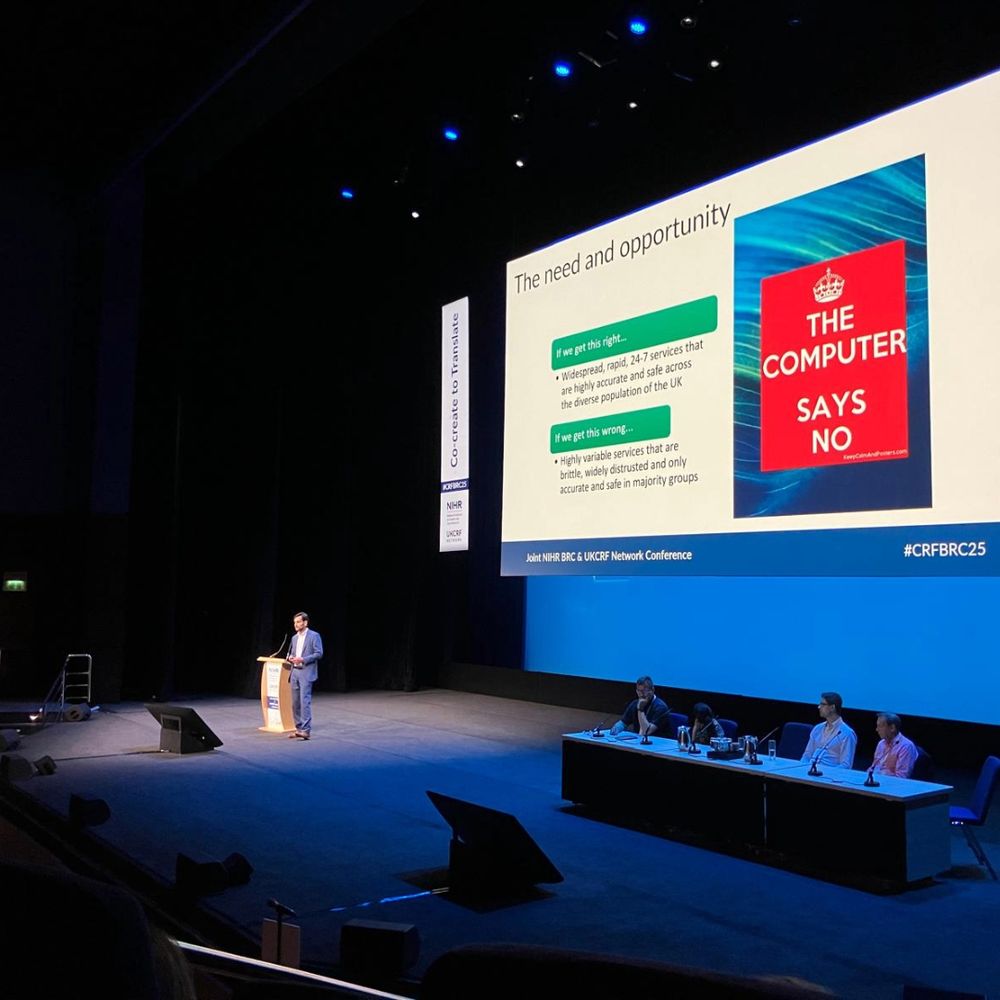  A man is standing at a podium on a stage, delivering a presentation at the CRF BRC & UKSPF Network Conference. Behind him, a large screen displays a slide titled 'The need and opportunity' with two bullet points discussing the consequences of getting data analysis right or wrong. An image of a red book cover titled 'THE COMPUTER SAYS NO' is also shown. Four people are seated at a table to the right of the stage.