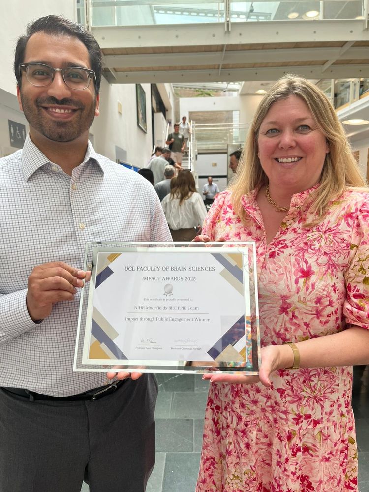 Two individuals standing indoors holding a framed certificate that reads 'UCL Faculty of Brain Sciences Impact Awards 2022 – Awarded to NIHR Moorfields BRC PPI Team – Impact through Public Engagement Winner.' The background features a modern building interior with people and stairs. 