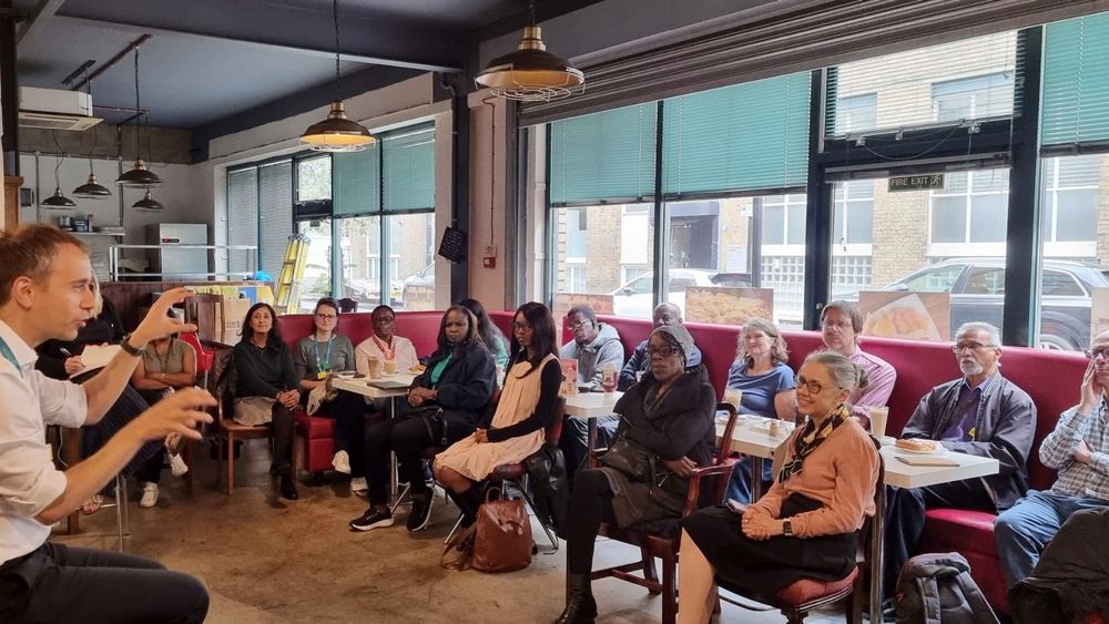 A group of people is seated in a room with large windows and red cushioned seating. The room features industrial-style pendant lights hanging from the ceiling. The group is facing a person who appears to be speaking or presenting, with hands gesturing. Tables in front of the seated individuals hold plates and cups. Outside the windows, buildings and parked cars are visible.