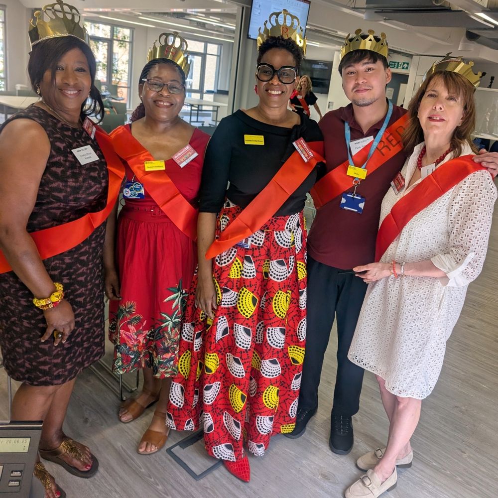 A group of five people standing together in a well-lit room. Each person is wearing a gold crown and a red sash across their chest. They are dressed in various outfits, including dresses and casual wear. The background shows an office-like environment with large windows and some furniture.