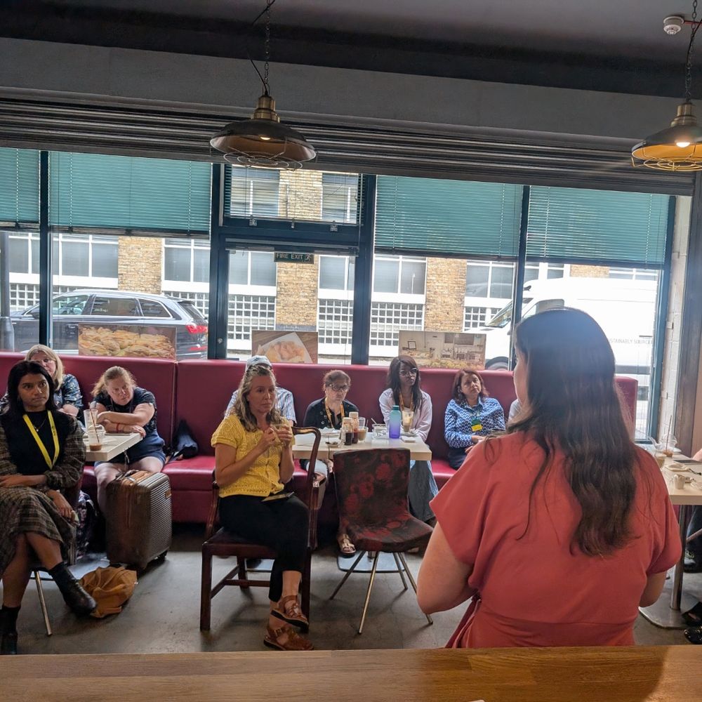 A group of people are seated in a cafe, facing a person standing at the front. The cafe has large windows with green blinds and brick buildings visible outside. The seated individuals appear to be engaged in an activity or discussion, with some holding notebooks or devices. The person standing has long hair and is wearing a red top.