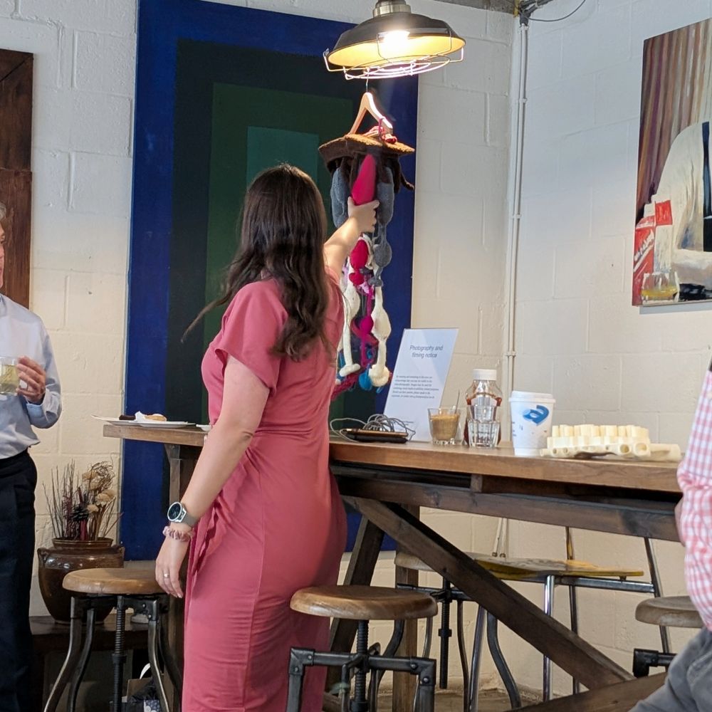 A group of people are seated in a cafe, facing a person standing at the front. The cafe has large windows with green blinds and brick buildings visible outside. The seated individuals appear to be engaged in an activity or discussion, with some holding notebooks or devices. The person standing has long hair and is wearing a red top. 