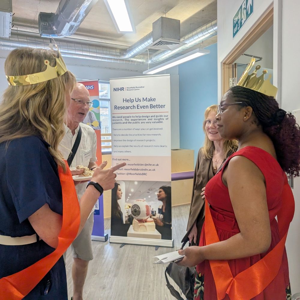 A group of people are standing and talking in a room. Two women are wearing gold crowns and orange sashes. In the background, a banner reads 'Help Us Make Research Even Better' and includes contact information for the NIHR, encouraging public involvement in research.