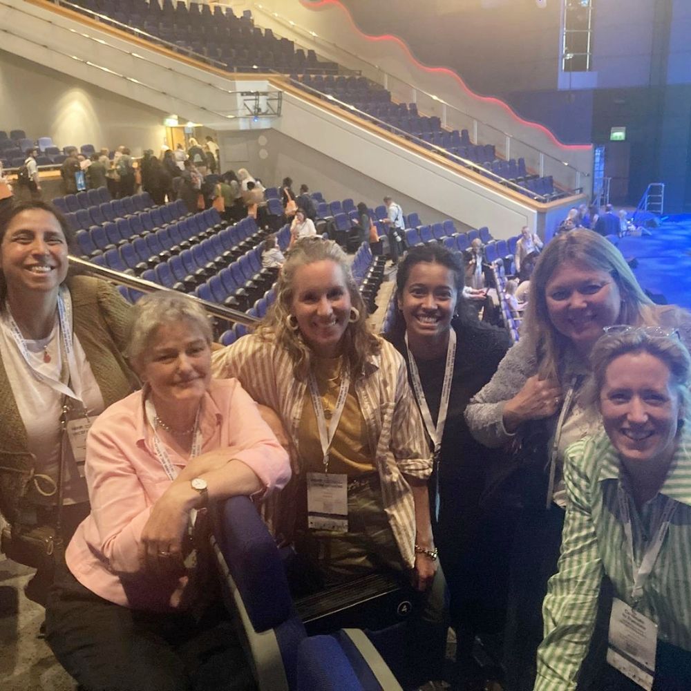  A group of five people standing and sitting in an auditorium with tiered seating. The auditorium has blue seats and is partially filled with other attendees. The people in the foreground are wearing conference badges around their necks, indicating they are attending an event or conference. The background shows more attendees mingling and walking around.