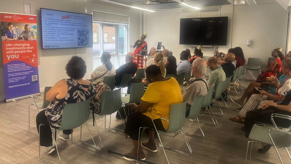 A group of people are seated in a room, facing a large screen displaying an agenda. A person stands at the front, possibly giving a presentation. On the left side, a banner promotes life-changing treatments and encourages participation in research.