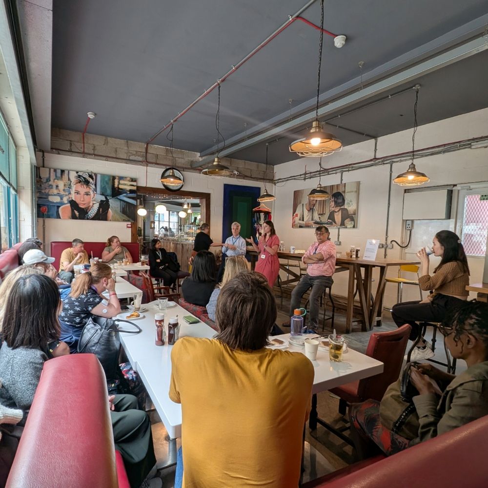 A group of people are seated in a cafe, facing a person standing at the front. The cafe has large windows with green blinds and brick buildings visible outside. The seated individuals appear to be engaged in an activity or discussion, with some holding notebooks or devices. The person standing has long hair and is wearing a red top. 