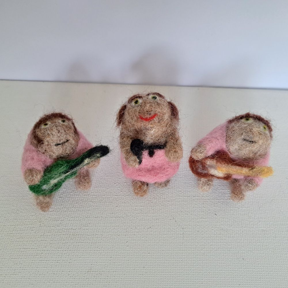 Three needle felted haggises dressed as the band Bis. The one on the left is John Disco with brown hair, sideburns, a pink shirt, and a dark green guitar. The one in the middle is Manda Rin with a pink and black dress, brown hair with bleach blonde streaks, white hair clips, and pinkish red lipstick. The one on the right is Sci Fi Steven with a pink shirt, brown hair, and reddish-brown and white guitar.