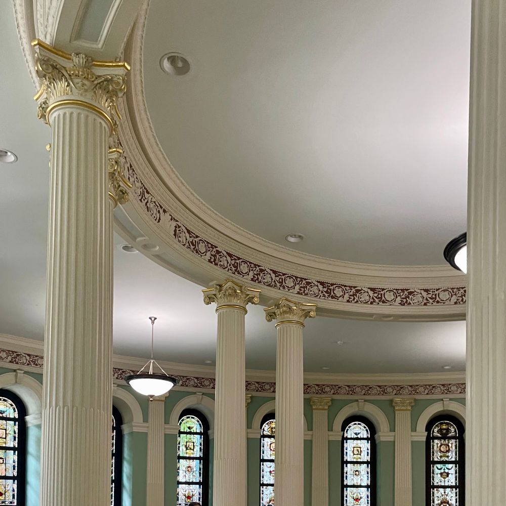 An interior showing an ornate classical rotunda-type set of pillars with gold leaf, there is mint green paint on the walls with stained glass windows. 