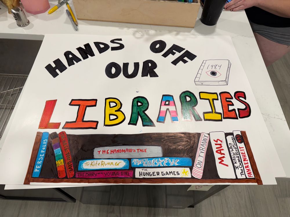 Protest sign “Hands off our libraries” with banned book titles on a bookshelf