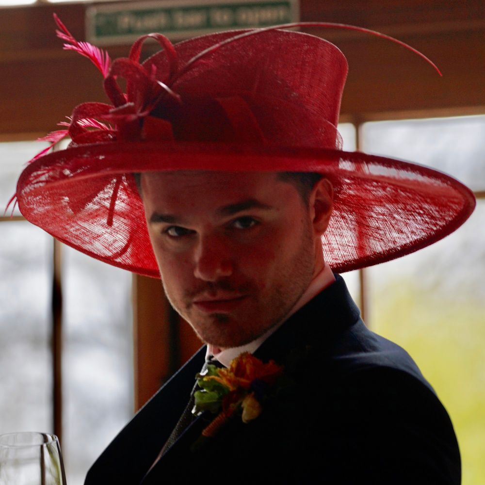 Isaac, wearing a navy wedding suit, a boutonnière and a colourful sinamay hat, pouting at the camera.
