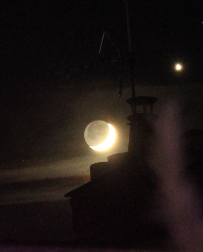 A dark night sky with a bright dot in the top right which I believe to be Venus in the centre is the moon which had a bright crescent to the bottom right quarter (about 1 - 7 on the clock face). There is the slight silhouette of a chimney in the foreground.