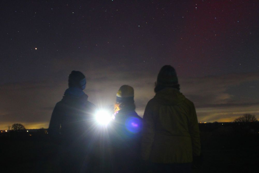 Faint red aurora borealis with three people silhouetted against the sky 