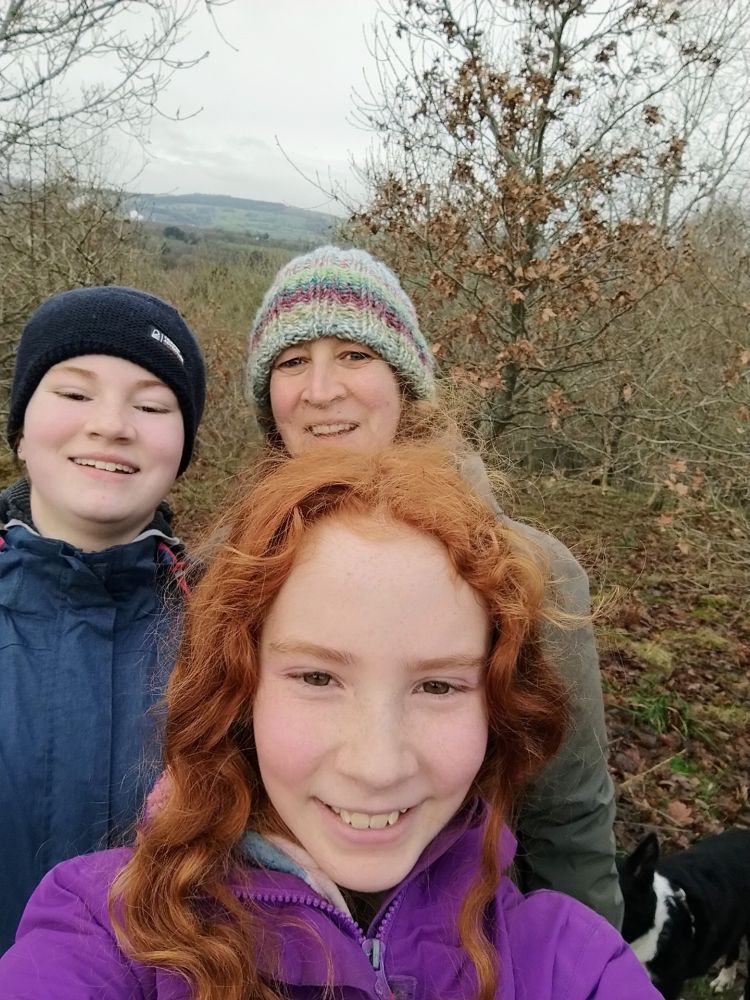 Grey sky and bare trees in the background behind a woman wearing a colourful knitted hat, a teen girl wearing a dark hat and blue waterproof coat, a younger girl with curly red hair wearing a purple waterproof coat. Everyone is smiling.