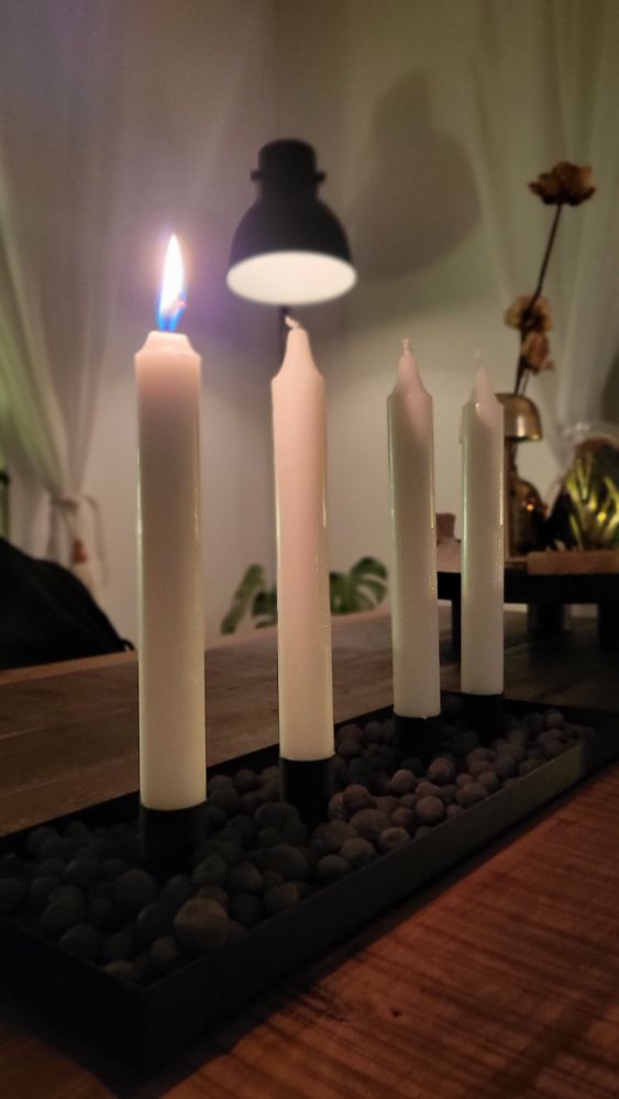 Fyra kronljus i en svart adventsljusstake, första ljuset brinner. Står på ett trädbord med en lampa i bakgrunden.