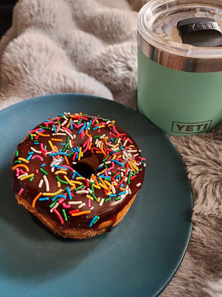 An expertly made chocolate dipped sprinkle doughnut on a blue plate rests on a snuggly warm grey fur blanketed lap with coffee in a teal Yeti mug.