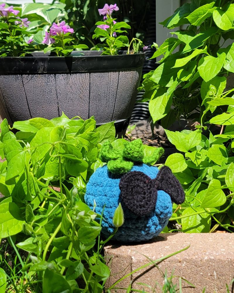 A blue qi fruit with sunglasses on in a garden with vibes around him and a basket of flowers behind. 