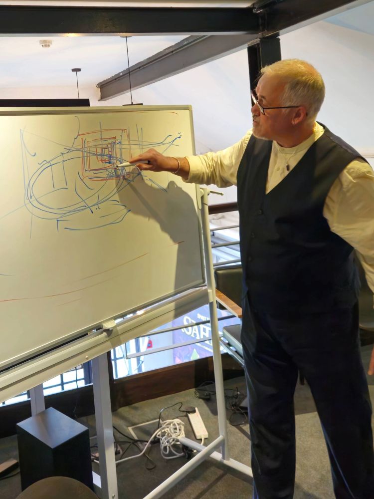 Francis Lowe, wearing glasses and a waistcoat, stands and points intently at a guide of Terry the Toucan flight, drawn in blue and red lines on a whiteboard in the studio.