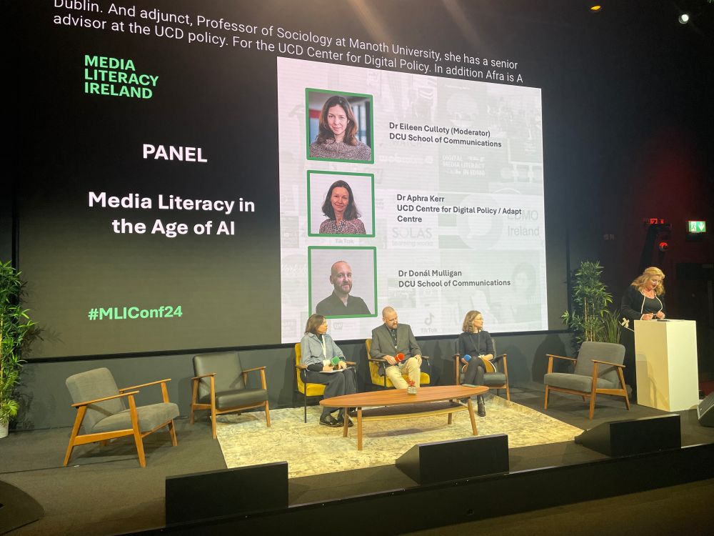 a panel discussion at the Media Literacy Ireland conference (#MLIConf24), focused on the topic "Media Literacy in the Age of AI". The panel features three experts:

    Dr. Eileen Culloty (Moderator, DCU School of Communications)
    Dr. Aphra Kerr (UCD Centre for Digital Policy / ADAPT Centre)
    Dr. Donal Mulligan (DCU School of Communications)

The stage setup includes modern seating, plants for a natural touch, and a large screen displaying the panel details and participant photos. A speaker, Martina Chapman, is addressing the audience from a podium. This event highlights critical discussions on navigating media literacy amidst the growing influence of artificial intelligence.