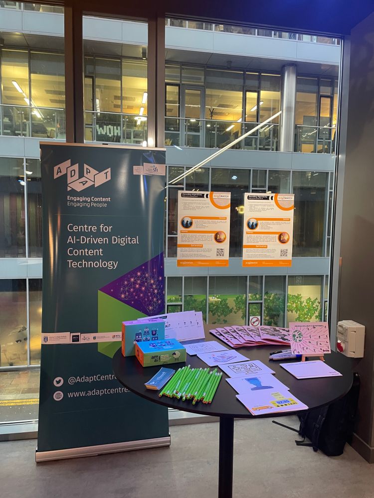 a display table at an event, set up by the Education & Public Engagement team from ADAPT Centre. 

The table includes colorful educational materials, pencils, and printed handouts. A banner prominently displays the ADAPT Centre's branding, emphasizing "Engaging Content, Engaging People" and its focus on AI-driven digital content technology. 

The setting is Media Literacy Ireland conference which took place at The Foundry in Google HQ, with large glass windows overlooking an interior office environment. This setup aims to engage attendees with information about AI and its applications including the Algowatch EU project focused on AI and algorithmic literacy.