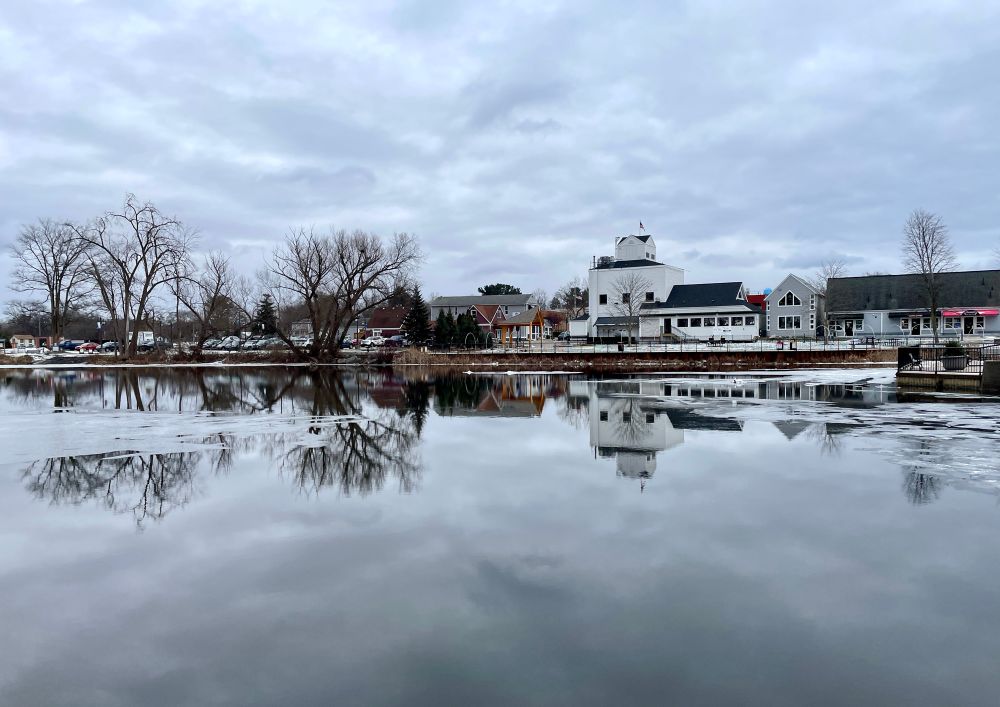 Photograph across a river of trees and buildings of a downtown.  It’s not a large city so buildings are smaller, more like a small town you would find in the countryside. The River is as smooth as glass, with small areas of ice. It’s a cloudy, overcast winter day and the clouds are reflected on the dark water. There is some snow on the ground around the buildings. The river shows a clear reflection of the trees, shrubs, and buildings that are on the opposite bank. 