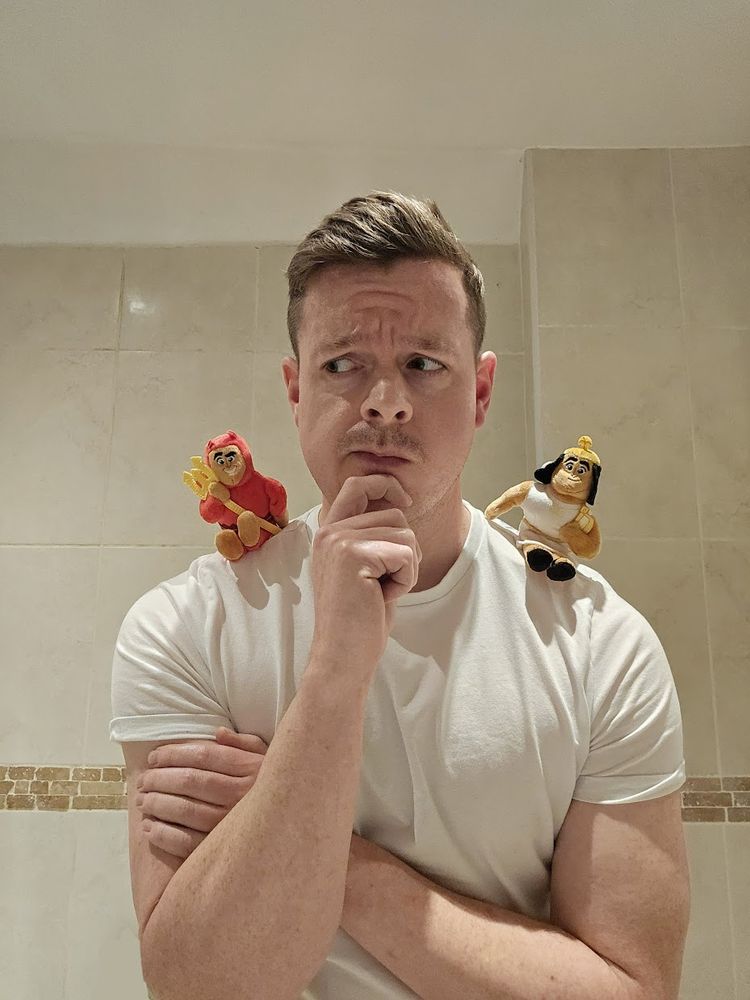 A man (me) looks quizzically with plush toy Kronk angel and devil figures on his shoulders
