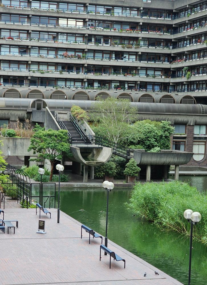 A large apartment block with a reflecting pool, dotted with 1970s street lamps and benches.