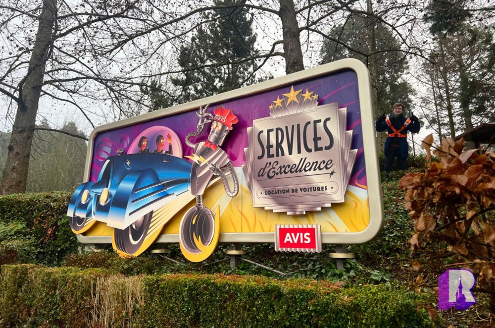 An Autopia billboard. On the flaming surface of a star, a robot in chauffeur's hat rolls alongside a retro-futuristic blue car. He tips his cap at a couple who wave back from the bubble dome of the vehicle. The Art Deco text reads "Services d'Excellence - Location de Voitures" "Premium Service, Car Rental" - Avis.