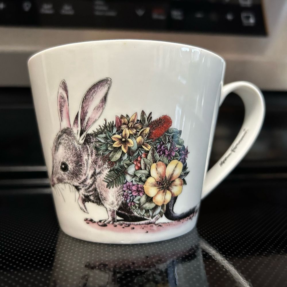 Maxwell & Williams Marini Ferlazzo 450 ml Australian Families Bilby Mug. The mug is white and wider at the top than at the base. There is a hand drawn bilby with flowers covering much of its body.