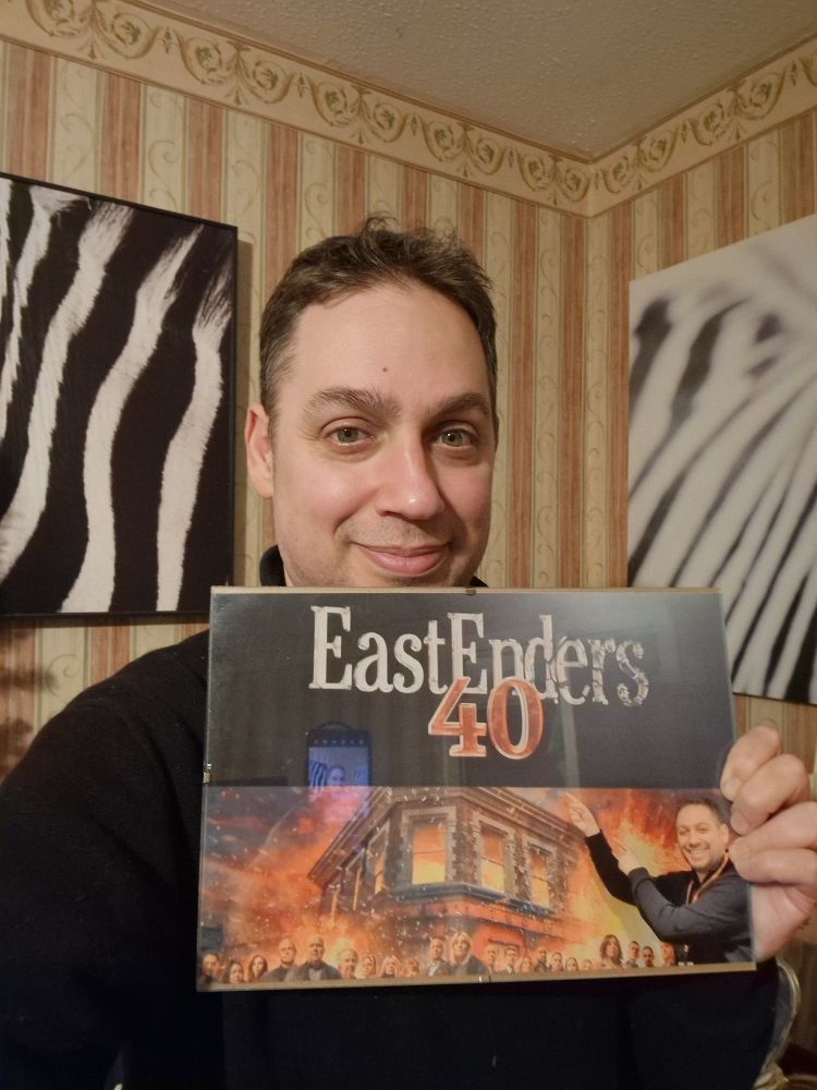Me holding an a4 photo reproduction, my mother gave me, of a photo I took at BBC Studios standing in front of an Eastenders 40th anniversary poster...