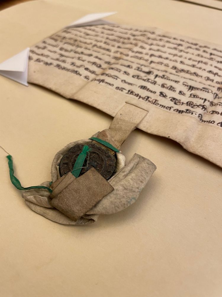 Medieval document with handwriting in black ink visible at top right, dark colored wax seal attached to bottom of document by a parchment strip, partially obscured by a cloth covering that has come loose, with two pieces of green ribbon once used to secure it.
