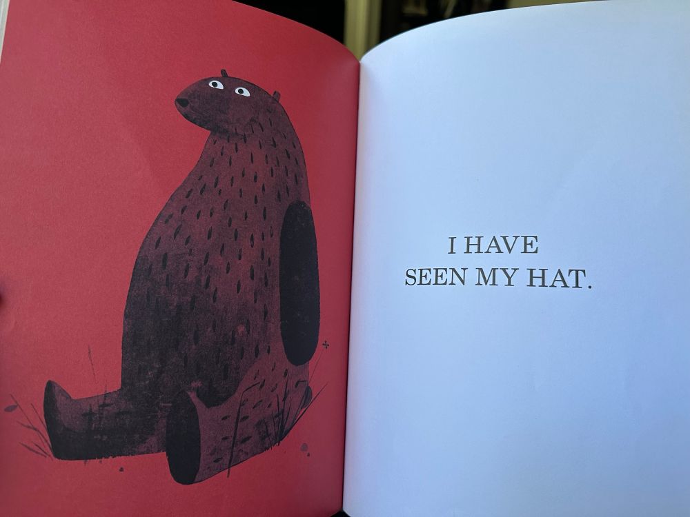 Two pages of an open children's book (I want my hat back, by Jon Klassen). The left page is red, with a picture of a seated bear with wide eyes. The right page is white and reads 