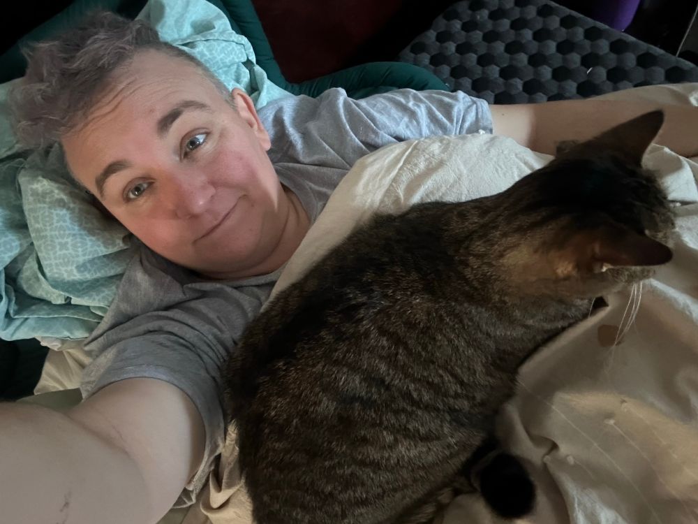 A vaguely smiling Caucasian woman with short grey hair lying against pillows as a brown and black tabby cat settles in on her chest.