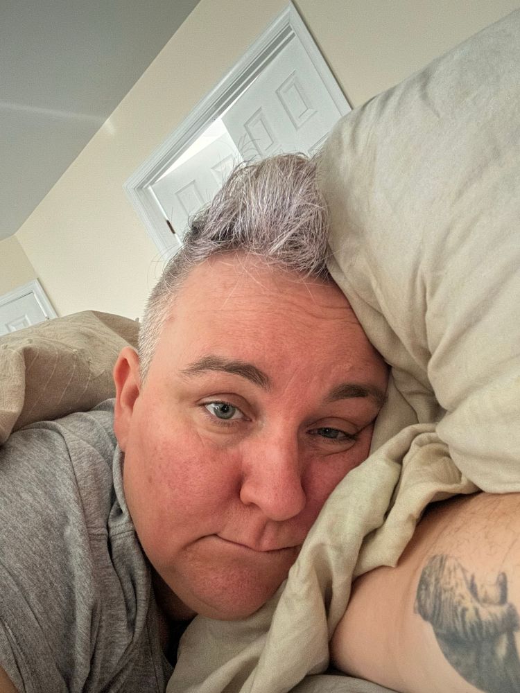 An exhausted looking woman (me!) with short grey hair lying in bed wondering whether the meat tenderizing hammer would work on her neck muscles.