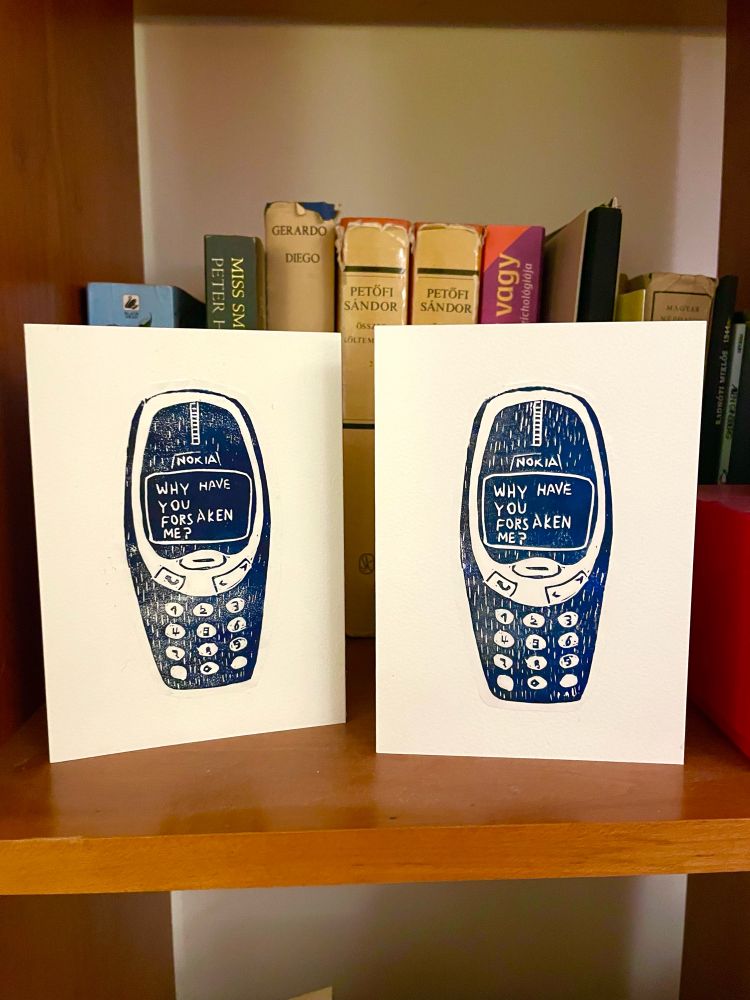 Two lino-printed cards (a Nokia mobile phone with the screen reading ‘why have you forsaken me?’) on a shelf with books 