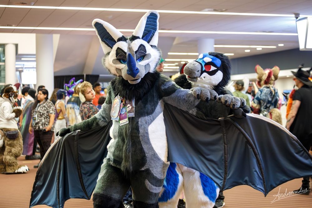 A bat fursuit with wings spread in front of a crowded hallway with a raptor fursuit peeking over the left wing. 