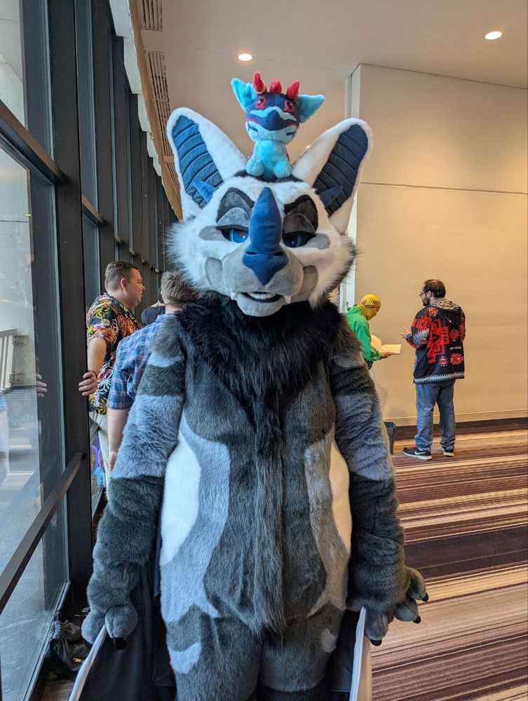 A very hungry bat fursuit with a small, plushie friend on top of his head. 