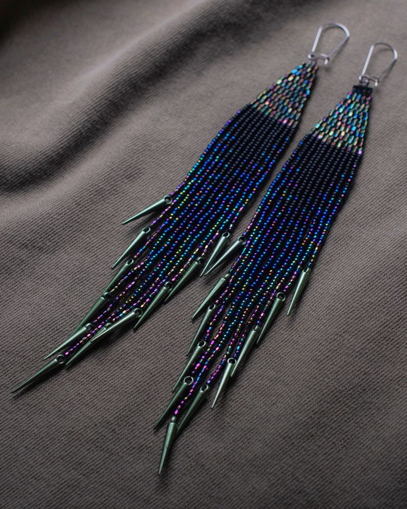 beaded fringe earrings. top triangle is a green iridescent hue. below the triangle it blends from black, blue, green, to purple. with ebony spikes. on a brown backdrop.