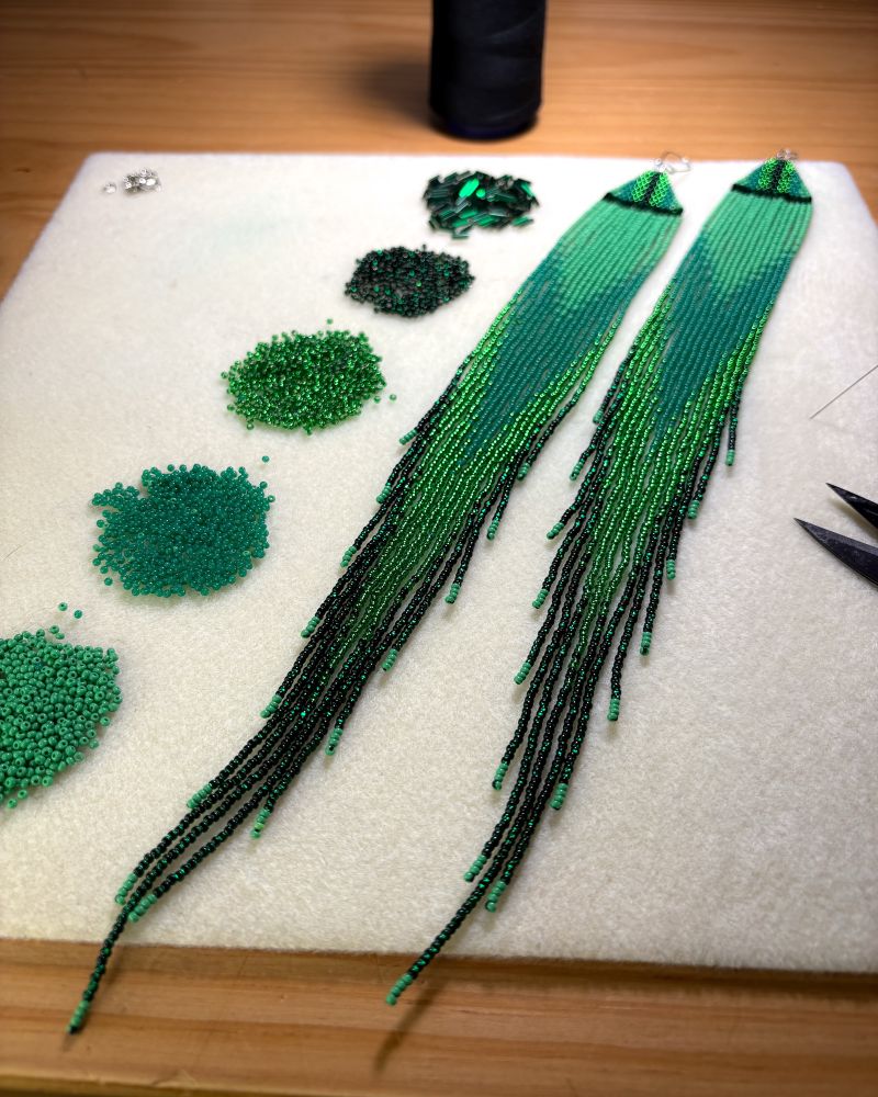 Large green beaded fringe earrings. A blend of light green to dark shiny green. On a bead mat with piles of beads on the left side laid out in a gradient from dark to light green. Black thread spool in the back. Thread needle & snippets poking in the photo on the right. 