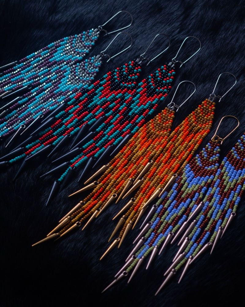 Colourful beaded fringe earrings with spikes at the ends on a black background. 1st pair represents water flow (crystal, baby blue, & purple) 2nd pair represents soapberries (black, red, teal) 3rd pair represents the sun on land (orange, dark brown, mustard) 4th pair represents the land (brown, powder blue, green)